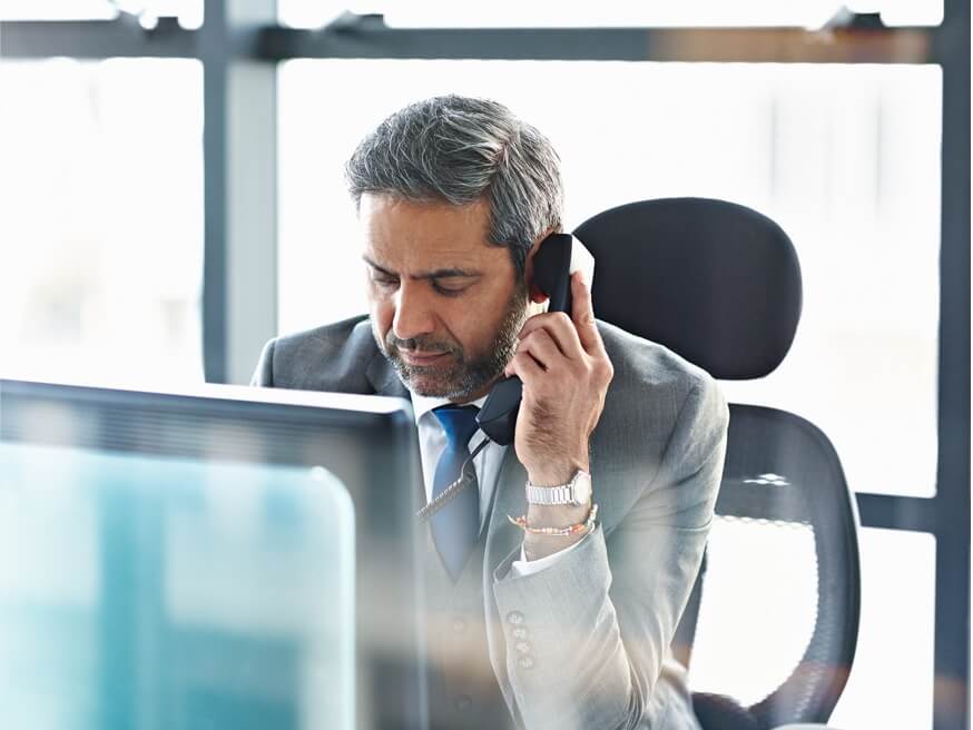 Man speaking on a desk phone, sitting in front of a monitor.